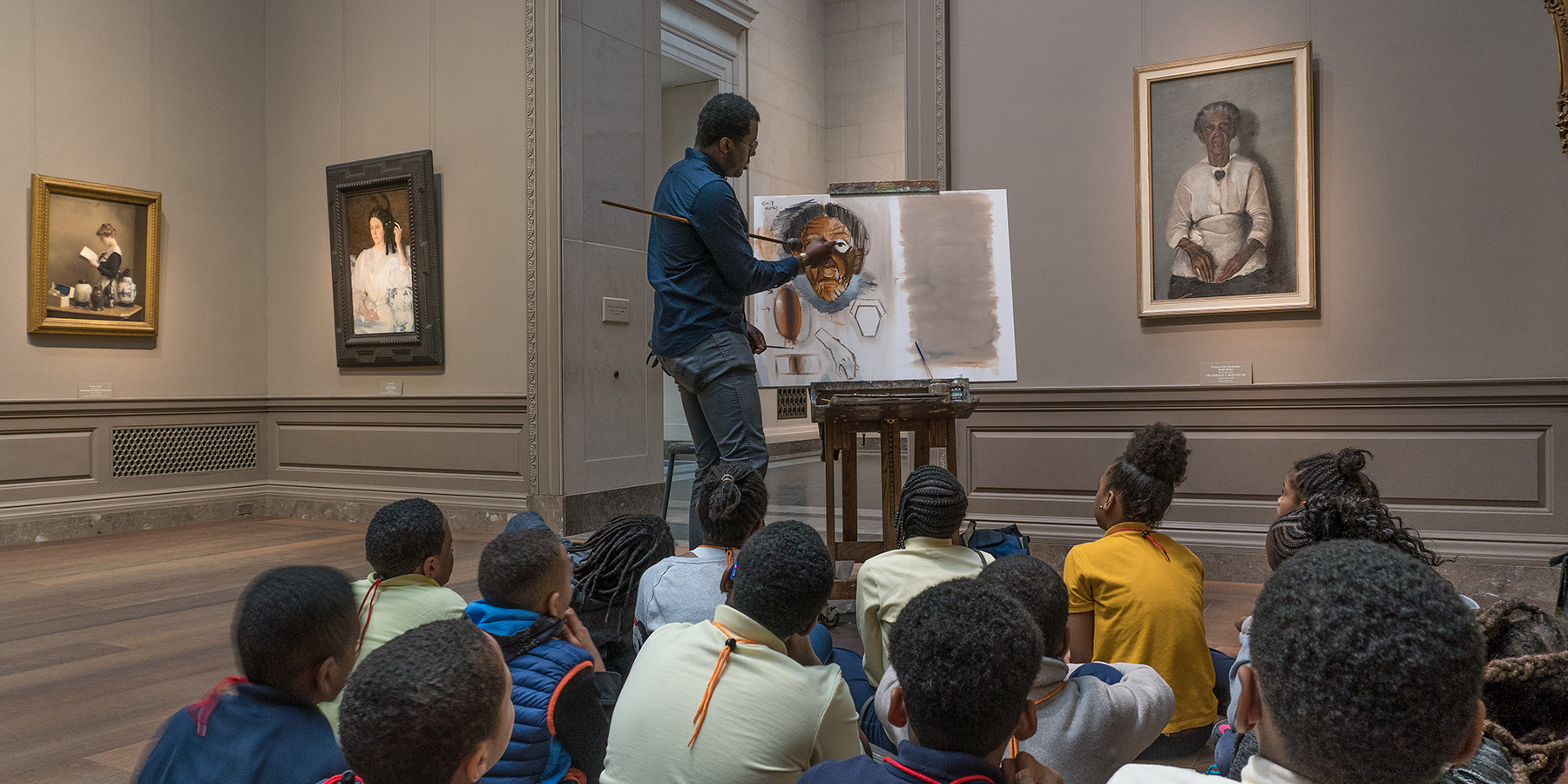 A man is painting a copy in the National Gallery of a painting in our collection as elementary school children are gathered around him watching him work.