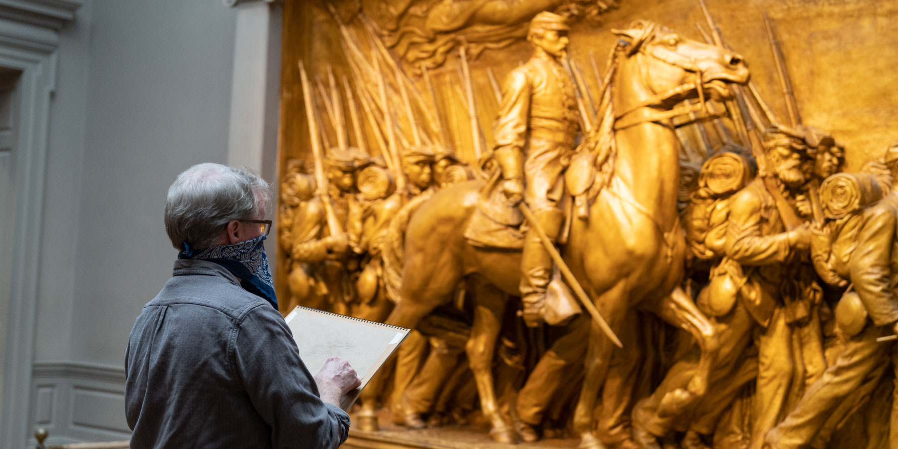 A person with a bandana facemask on is sketching a sculpture depicting Union Army soldiers marching off to battle with their officer on his horse.