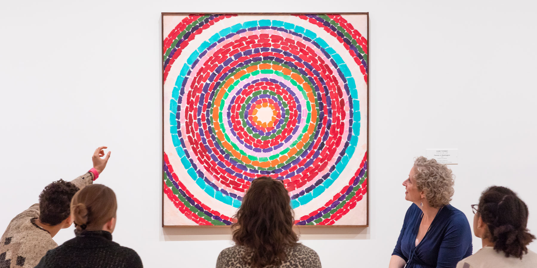 Five people are seated in a semi-circle looking at a painting that shows many multicolored circles radiating out from the middle of the composition. One of the people is pointing at the painting. 