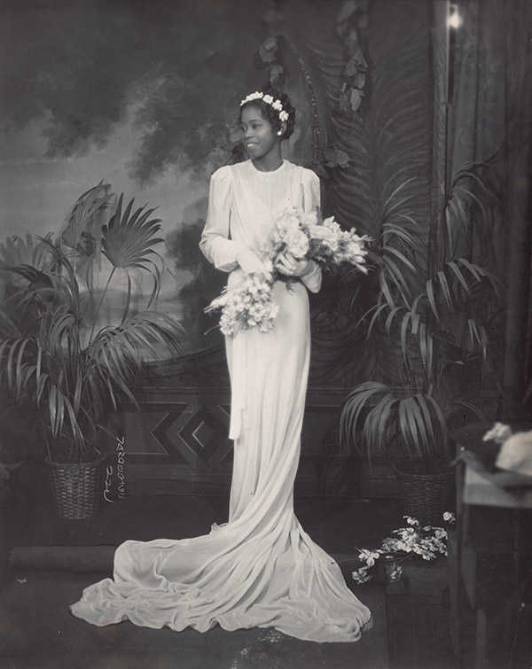 A woman in white wedding dress wearing a white flower headband holding a bouquet of flowers with palm plants behind her