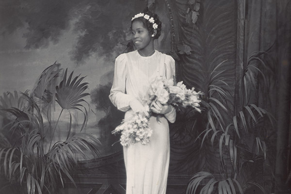 A woman in white wedding dress wearing a white flower headband holding a bouquet of flowers with palm plants behind her
