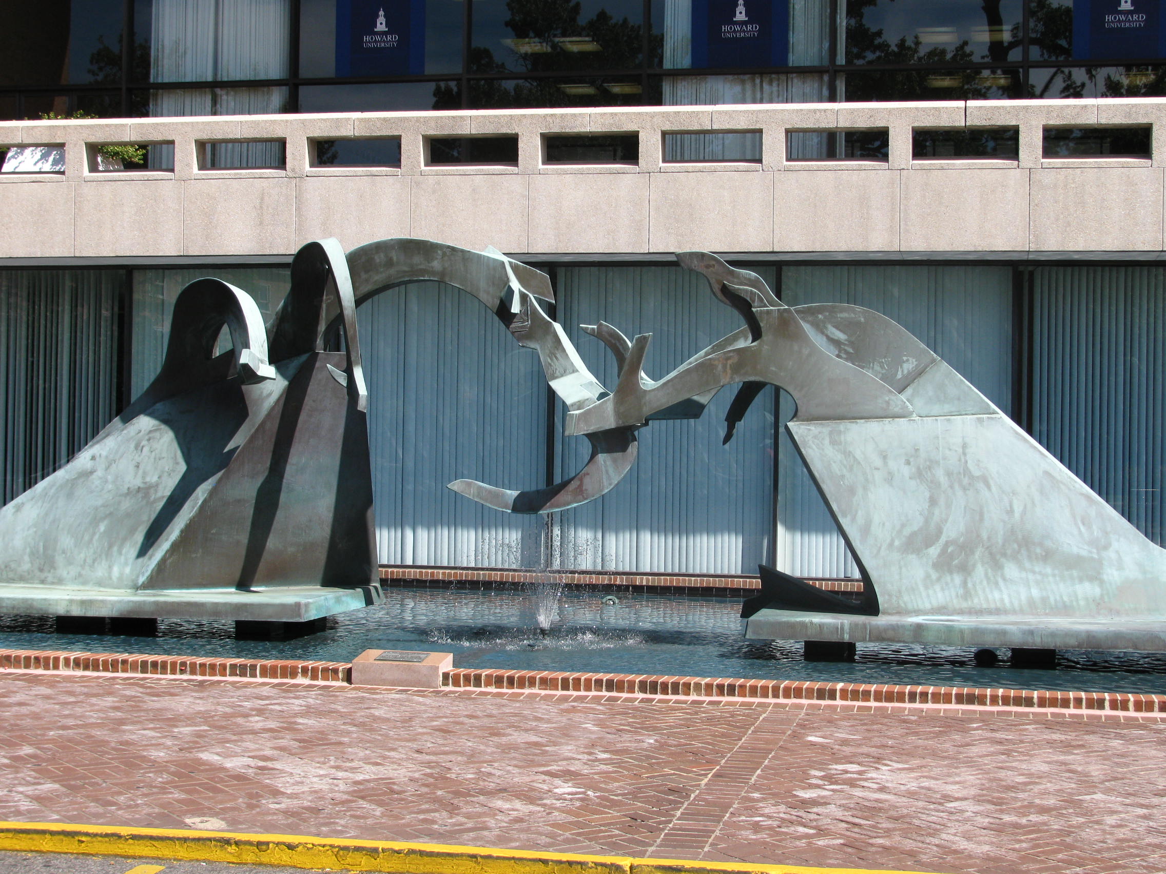 A metalic sculpture sits outside over a rectangual water fountain in front of office windows. The sculpture rises up and interlocks between the left side and right side in abstract shapes.