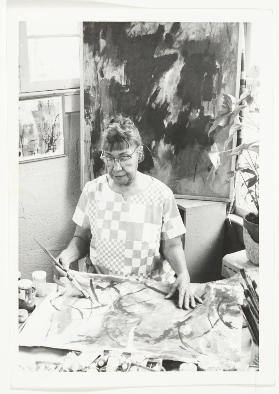 Black-and-white photograph of woman painting in a studio surrounded by art supplies, painted canvases, and a plant.