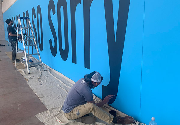 Two men painting SO-SO, SO SO, SORRY on a blue blackground installed across the front of the entrance East Building of the National Gallery of Art