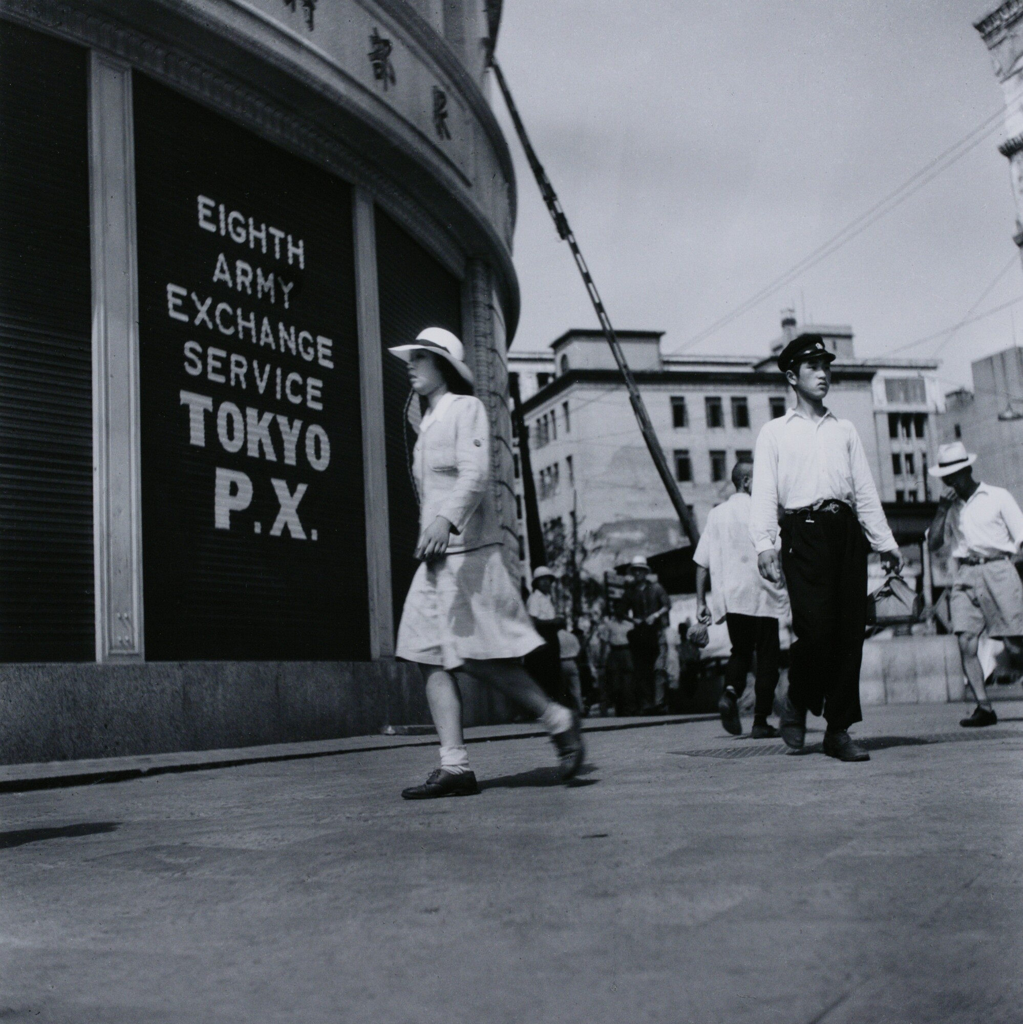 A woman walking down city street right to left with light-colored hat, jacket, and dress. Three men behind her on the right of the composition looking in different directions. The first man is standing looking off to the right in a dark hat, light shirt, and dark pants. Another man is behind him on the right looking down with a light-colored hat, shirt, and shorts. Another man is behind the first man to the left in a light-colored shirt and dark pants. City buildings in the background with other people in the distance.