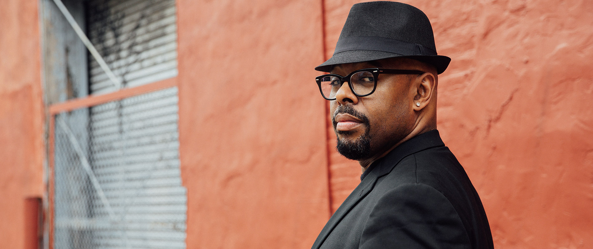 A man in a black suit, wearing a black fedora hat, black rimmed glasses and dark goatee looking back at the viewer over his left shoulder. An orange wall is in the background along with a gray garage door off into the distance on the left side of the composition.