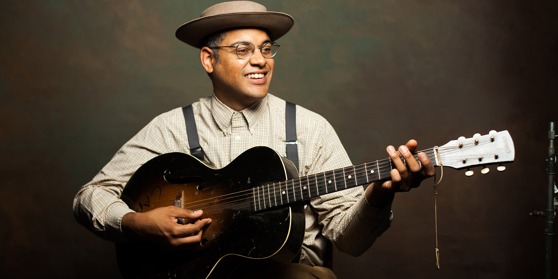 Man with an old-timey hat, round glasses, suspenders, button-up long-sleeve shirt playing a hollow-body guitar with f-holes