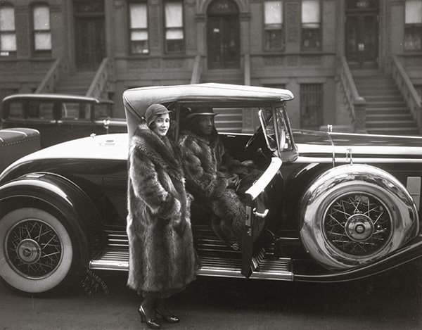 A man in a fur coat and fedora sitting in a parked car from the 1930s with door open and a woman standing next to him wearing a fur coat and flapper-style hat.