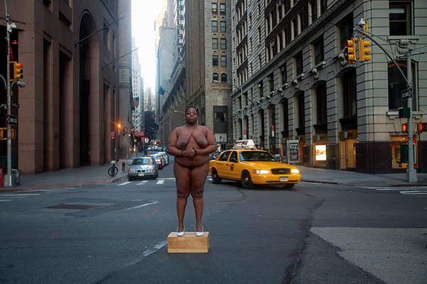 A Black woman stands on a box on a city street and stares into the camera. She is not clothed