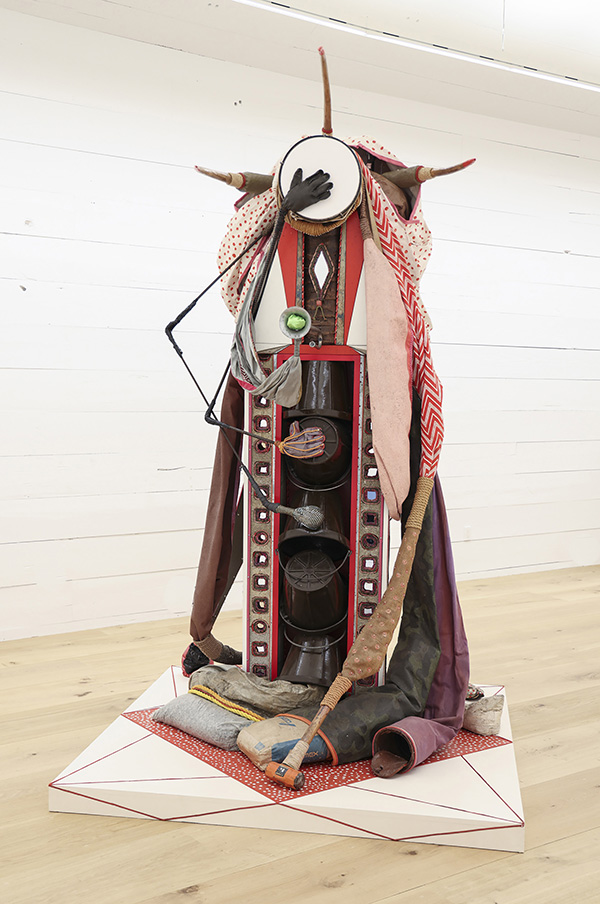 This is a photograph of a sculpture. It's a person like form with a drum for a head and a stick arm with a glove that covers the face. The figure is draped in fabrics