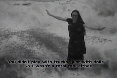 Womanon the beach wearing a black dress as a wave crashes in on her legs with closed captioned "You didn't play with trucks just with dolls. So I wasn't a total fool either."