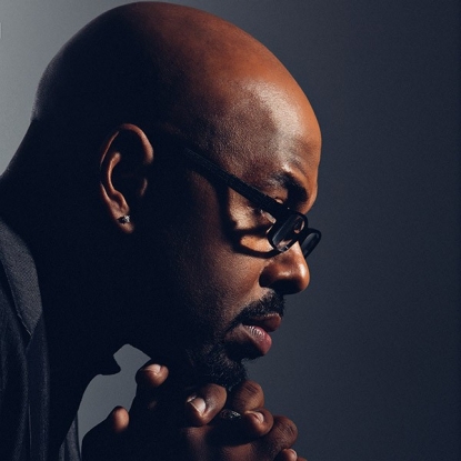 A profile of a man's face looking to the viewers right and straight ahead from his perspective. His fingers are interlocked in a prayer-like pose and are resting below his face on his chin. He is completely bald, has black-rimmed glasses, and a goatee. 