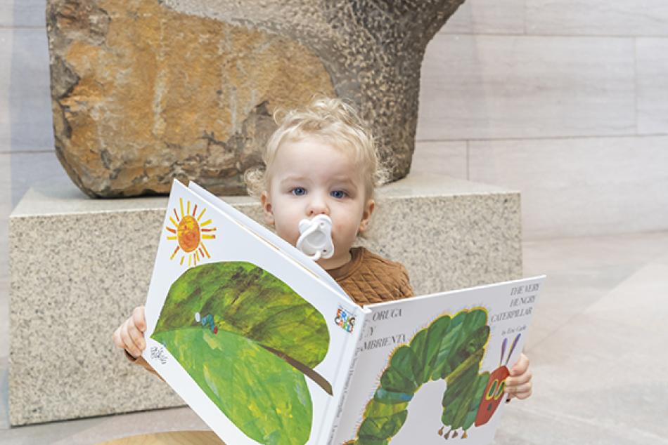 A very young visitor reading a children's book with a pacifier in it's mouth.