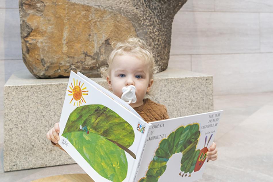 A very young visitor reading a children's book with a pacifier in it's mouth.