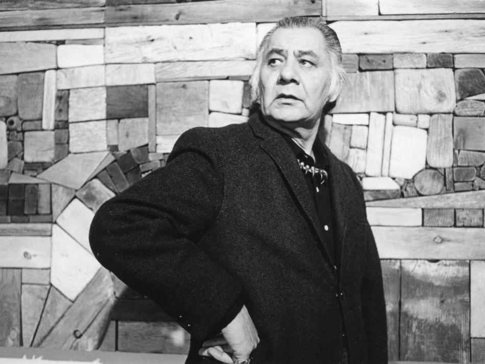 A black and white photograph shows a man with greyish hair standing against a wall of stacked cubes of wood in different shades. He is shown from the waist up. He is turned to the right and looks to the left. He rests his left hand on his waist, with his elbow bent. He has a serious expression.