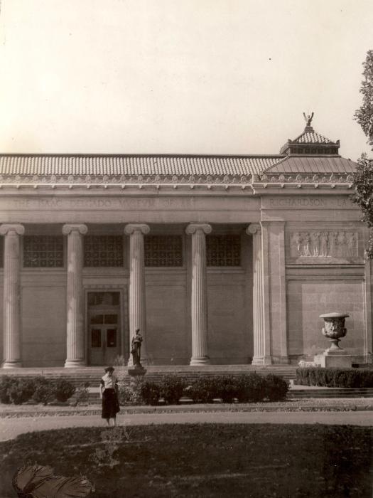 A black and white image of a neoclassical building facade with a row of columns. A woman with dark hair and light skin stands in the foreground wearing a dress, looking down at a reflecting pond.
