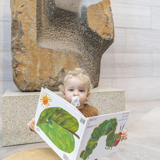 A very young visitor reading a children's book with a pacifier in it's mouth.