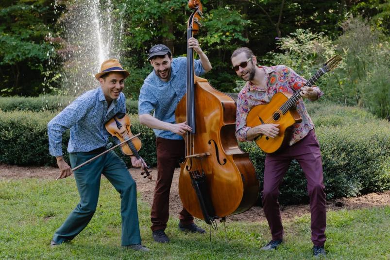Three men playing musical instruments in a park with a water fountain behind them.
