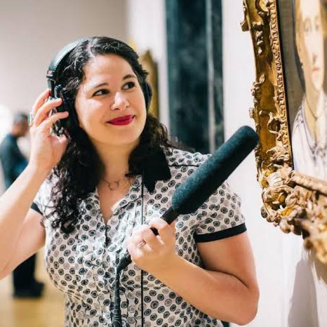Tamar Avishai wears headphones and holds a microphone up in front of a painting