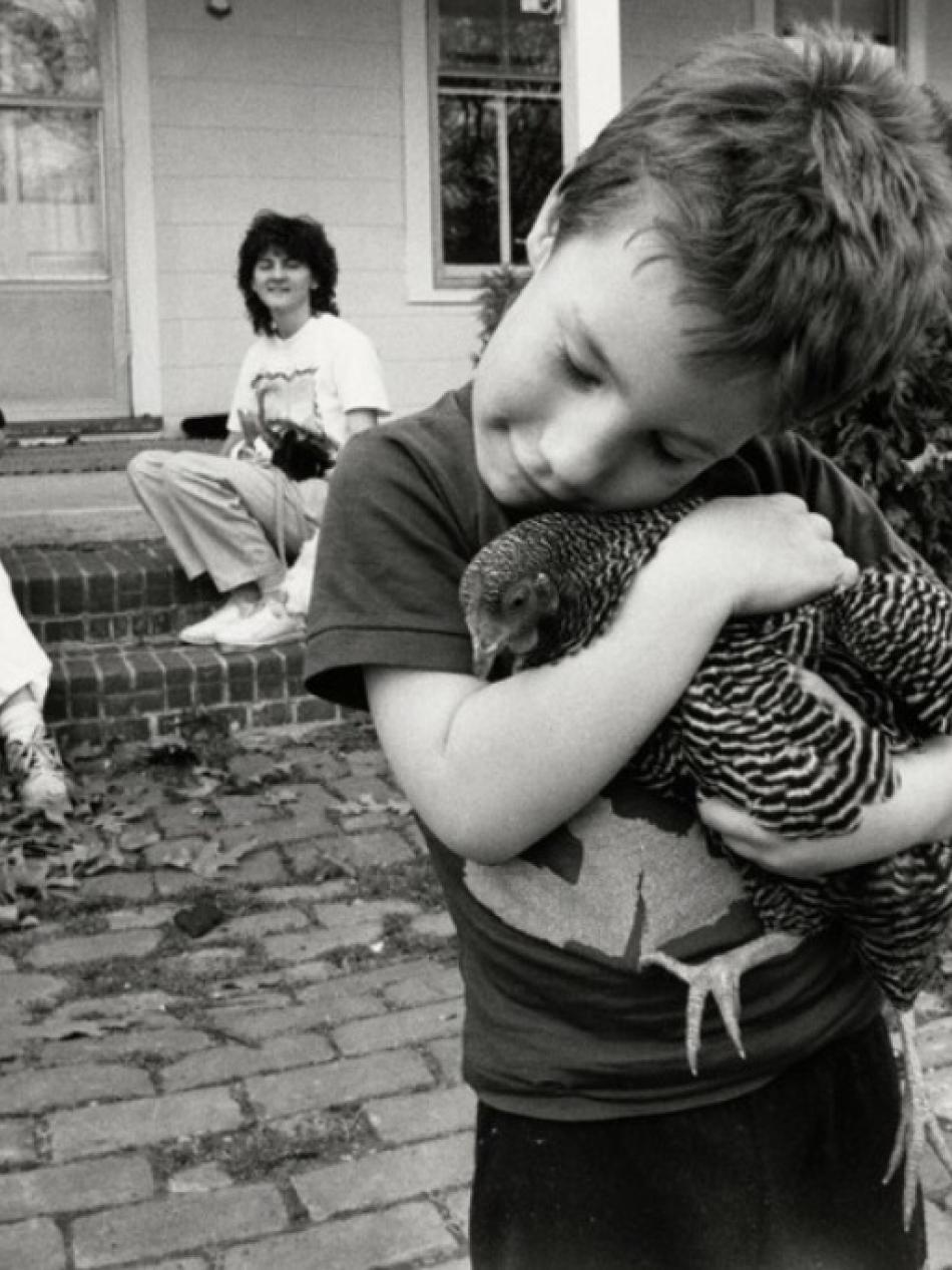 A young boy lovingly holds a chicken in this black and white photograph. Two women sit on the porch in the background.