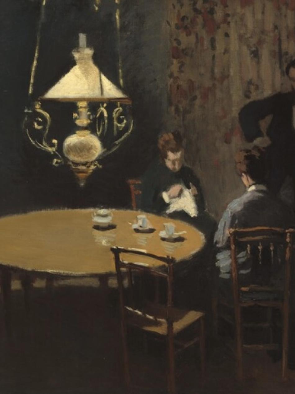 Two people sit at a table while one stands. There is a hanging light over the table.