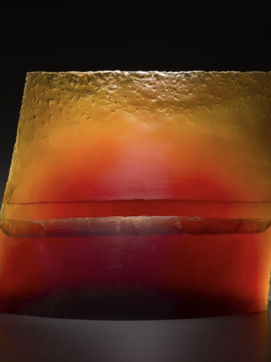 This vertical piece of glass is slightly tilted to the right at the top. It is orange an opaque at the bottom and more translucent at the top.