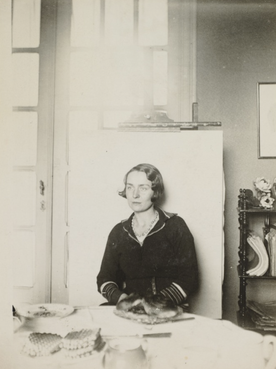 This is a black-and-white photograph of a woman seated at a table with food items, including waffles. She wears a dark dress with striped cuffs and a necklace, and sits in front of a window and a tall shelf with stacked papers and a vase.