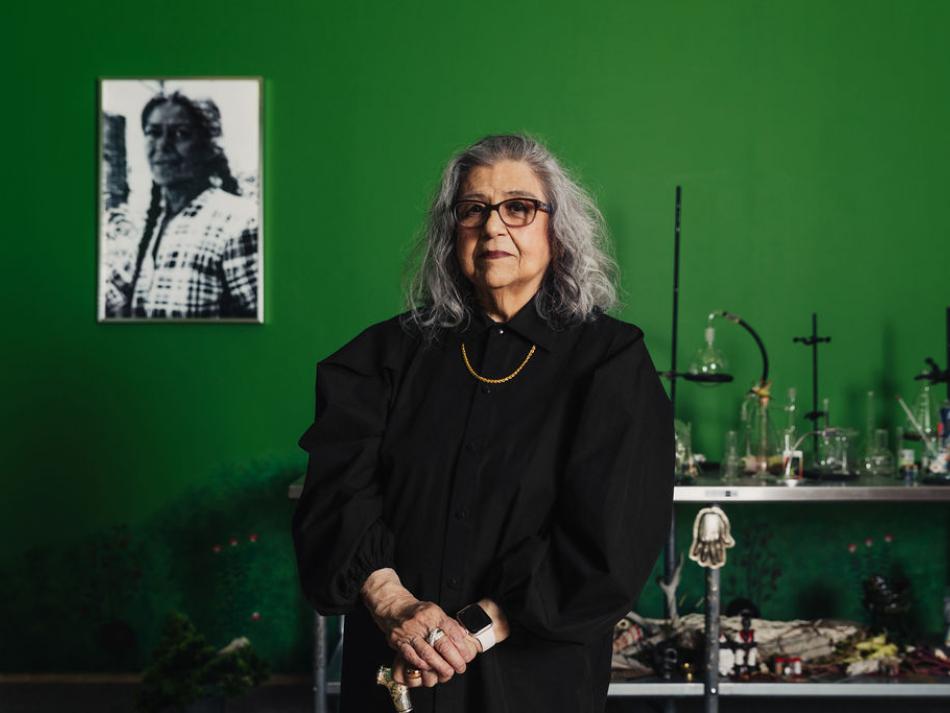 A woman with white grey hair and black glasses stands with her hands clasped at her waist. She is shown from the waist up. Behind her is a bright green wall, with a black and white photograph of a woman to the left. On her right is a metal table of various scientific tools and other small objects.